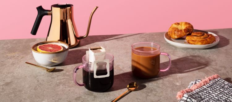 Photo of two cups of coffee, a bowl of grapefruit, spoons, and a plate of pastries on a marble table.